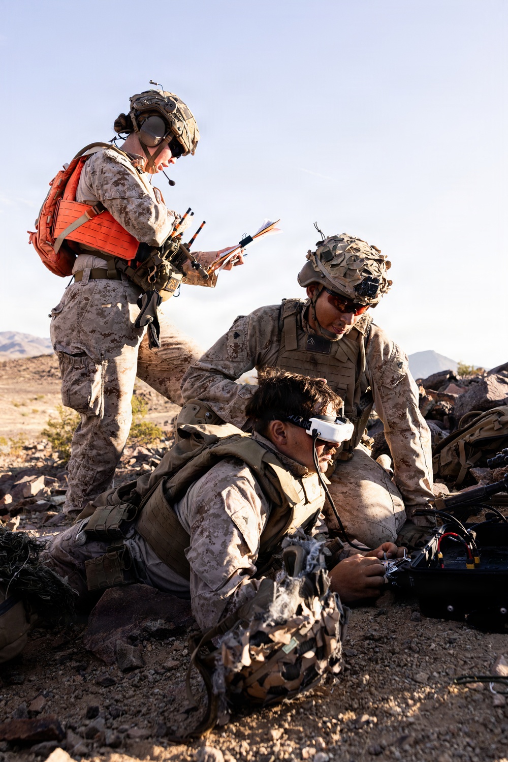 Three military personnel in MARPAT desert camouflage uniforms are operating in a rocky desert environment. One soldier wearing FPV (First Person View) goggles is prone on the ground handling what appears to be a drone controller or electronic device, while a second soldier assists, and a third standing soldier wearing a red safety vest and helmet with communications equipment reviews documents. The setting and equipment suggest a drone operations training exercise.