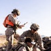 Three military personnel in MARPAT desert camouflage uniforms are operating in a rocky desert environment. One soldier wearing FPV (First Person View) goggles is prone on the ground handling what appears to be a drone controller or electronic device, while a second soldier assists, and a third standing soldier wearing a red safety vest and helmet with communications equipment reviews documents. The setting and equipment suggest a drone operations training exercise.