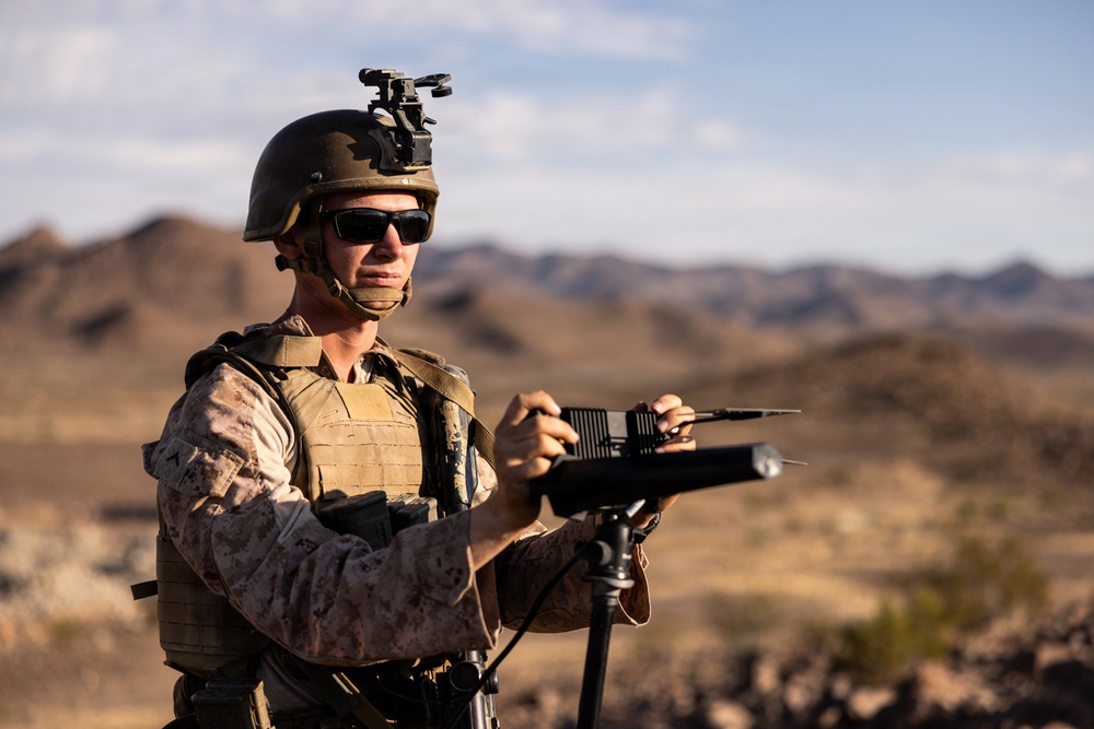 A military service member in desert camouflage uniform operates what appears to be a directional antenna or electronic warfare/communications device mounted on a tripod. The setting is an arid desert environment with mountains visible in the background. The soldier is equipped with full tactical gear including a combat helmet with night vision mount, plate carrier vest, and tactical eyewear.