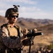 A military service member in desert camouflage uniform operates what appears to be a directional antenna or electronic warfare/communications device mounted on a tripod. The setting is an arid desert environment with mountains visible in the background. The soldier is equipped with full tactical gear including a combat helmet with night vision mount, plate carrier vest, and tactical eyewear.