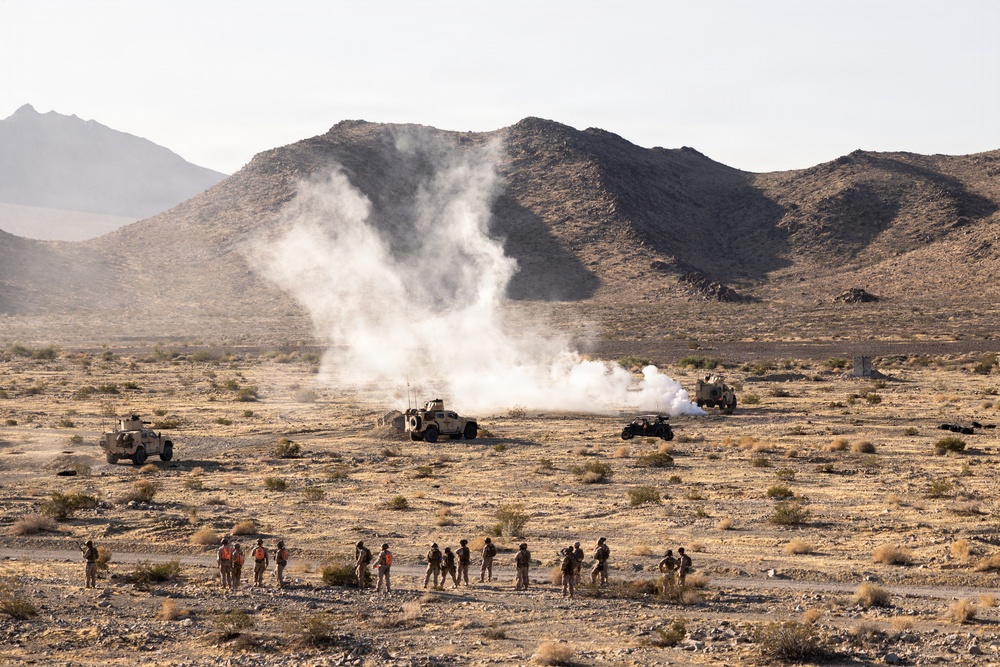 A military training exercise is taking place in an arid desert environment with rocky mountains in the background. Several military tactical vehicles are positioned in the mid-ground while a large smoke or explosion plume rises between them. A group of approximately 15-20 personnel, some wearing high-visibility orange vests, observe from a safe distance in the foreground.