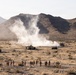A military training exercise is taking place in an arid desert environment with rocky mountains in the background. Several military tactical vehicles are positioned in the mid-ground while a large smoke or explosion plume rises between them. A group of approximately 15-20 personnel, some wearing high-visibility orange vests, observe from a safe distance in the foreground.