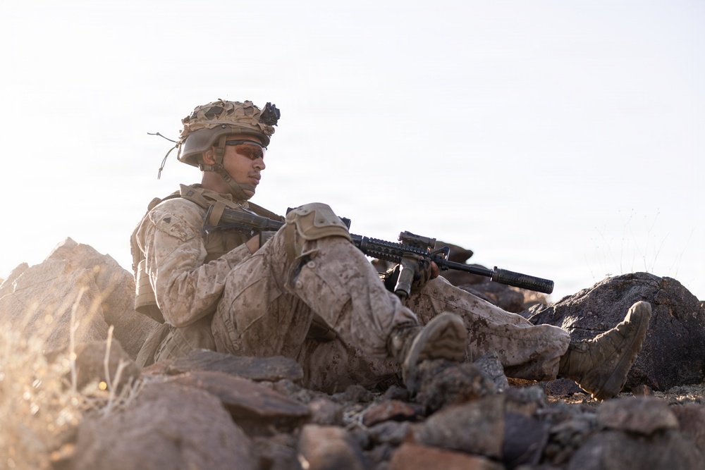 A soldier in desert camouflage (MARPAT pattern) is seated among rocks in an arid terrain, holding an assault rifle with an underbarrel attachment. The individual is wearing full combat gear including a tactical helmet with mounted equipment, face paint, and body armor. The setting appears to be a rocky desert environment photographed during golden hour lighting.