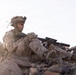 A soldier in desert camouflage (MARPAT pattern) is seated among rocks in an arid terrain, holding an assault rifle with an underbarrel attachment. The individual is wearing full combat gear including a tactical helmet with mounted equipment, face paint, and body armor. The setting appears to be a rocky desert environment photographed during golden hour lighting.