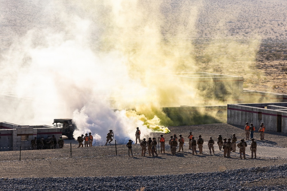 A military training exercise is underway at what appears to be a desert training facility with mock urban structures. A large yellow-green smoke grenade has been deployed, creating a significant smoke cloud over the training area. A group of uniformed personnel, some wearing orange safety vests, observe or participate in the exercise from a safe distance.
