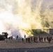 A military training exercise is underway at what appears to be a desert training facility with mock urban structures. A large yellow-green smoke grenade has been deployed, creating a significant smoke cloud over the training area. A group of uniformed personnel, some wearing orange safety vests, observe or participate in the exercise from a safe distance.
