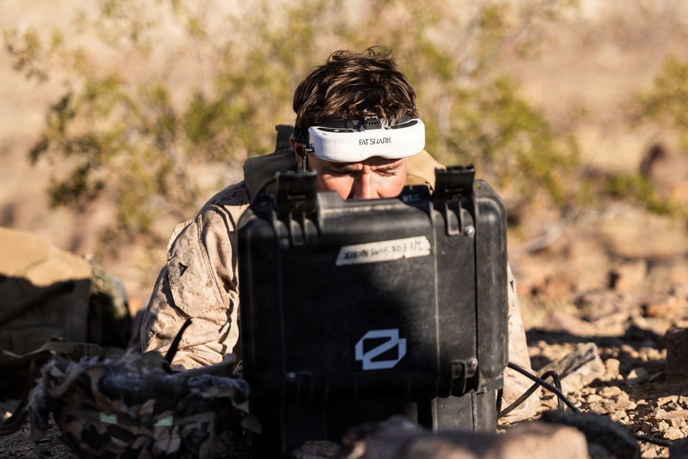 A person wearing desert camouflage military uniform and a white 'Fat Shark' FPV (First Person View) headset is operating equipment from behind a black hard-shell protective case in an arid, rocky outdoor environment. The individual appears to be engaged in drone or remote vehicle operation, using the FPV goggles pushed up on their forehead while looking into or over the equipment case. The setting suggests a military training exercise or field operation.