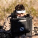 A person wearing desert camouflage military uniform and a white 'Fat Shark' FPV (First Person View) headset is operating equipment from behind a black hard-shell protective case in an arid, rocky outdoor environment. The individual appears to be engaged in drone or remote vehicle operation, using the FPV goggles pushed up on their forehead while looking into or over the equipment case. The setting suggests a military training exercise or field operation.