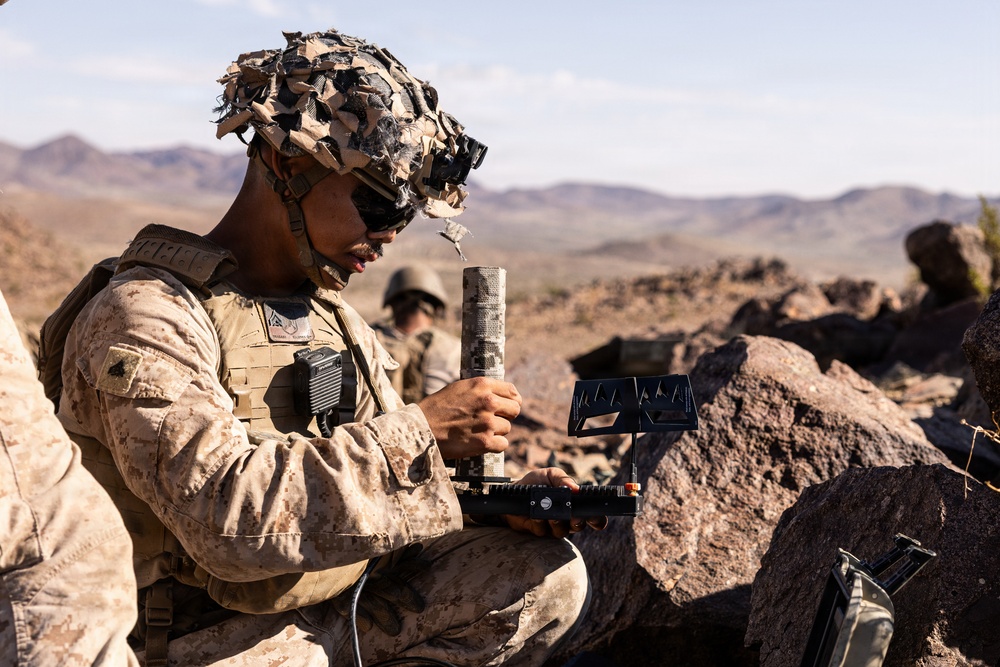A U.S. Marine in desert MARPAT camouflage uniform and a camouflage-covered helmet is operating what appears to be a drone ground control station or electronic warfare device in a rocky desert environment. A second service member is visible in the background. The setting appears to be a training exercise in an arid, mountainous desert region.