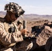 A U.S. Marine in desert MARPAT camouflage uniform and a camouflage-covered helmet is operating what appears to be a drone ground control station or electronic warfare device in a rocky desert environment. A second service member is visible in the background. The setting appears to be a training exercise in an arid, mountainous desert region.