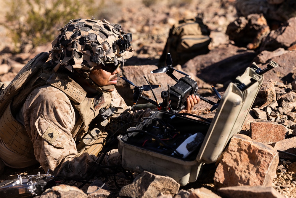 A military service member in desert camouflage uniform and a camouflage-covered helmet is lying prone on rocky terrain while handling a small quadcopter drone. The soldier is working with the drone over an open hard-sided carrying case, suggesting preparation for deployment or post-mission recovery. The setting appears to be an arid, rocky desert environment consistent with a training or operational scenario.