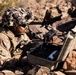 A military service member in desert camouflage uniform and a camouflage-covered helmet is lying prone on rocky terrain while handling a small quadcopter drone. The soldier is working with the drone over an open hard-sided carrying case, suggesting preparation for deployment or post-mission recovery. The setting appears to be an arid, rocky desert environment consistent with a training or operational scenario.