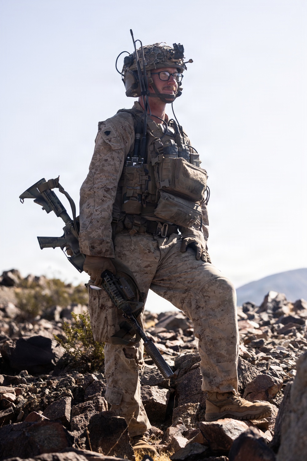 A fully equipped military service member stands on rocky terrain in what appears to be a desert environment. The individual is wearing USMC MARPAT desert camouflage and carrying extensive tactical gear including communications equipment and a rifle. The setting suggests a field training exercise or operational deployment in an arid, mountainous region.