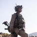 A fully equipped military service member stands on rocky terrain in what appears to be a desert environment. The individual is wearing USMC MARPAT desert camouflage and carrying extensive tactical gear including communications equipment and a rifle. The setting suggests a field training exercise or operational deployment in an arid, mountainous region.