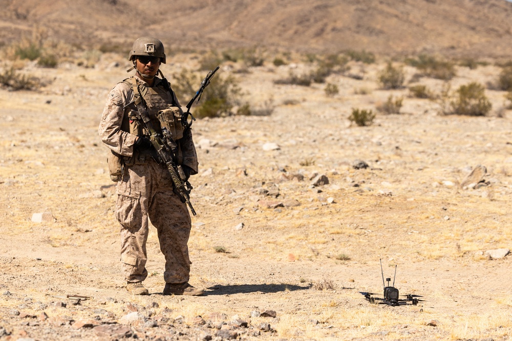 A uniformed military service member stands in an arid desert environment, equipped with full combat gear including a rifle and tactical vest. A small quadcopter drone is visible on the ground to the right of the soldier. The setting appears to be a training exercise in a desert region, likely in the American Southwest based on the terrain and vegetation.