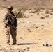 A uniformed military service member stands in an arid desert environment, equipped with full combat gear including a rifle and tactical vest. A small quadcopter drone is visible on the ground to the right of the soldier. The setting appears to be a training exercise in a desert region, likely in the American Southwest based on the terrain and vegetation.