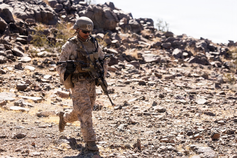 A single soldier in desert camouflage uniform is patrolling on foot through a rocky, arid terrain. The individual is fully equipped with combat gear including a helmet, body armor, tactical vest, and an assault rifle. The environment appears to be a desert landscape with scattered rocks and sparse dry vegetation.