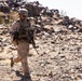 A single soldier in desert camouflage uniform is patrolling on foot through a rocky, arid terrain. The individual is fully equipped with combat gear including a helmet, body armor, tactical vest, and an assault rifle. The environment appears to be a desert landscape with scattered rocks and sparse dry vegetation.