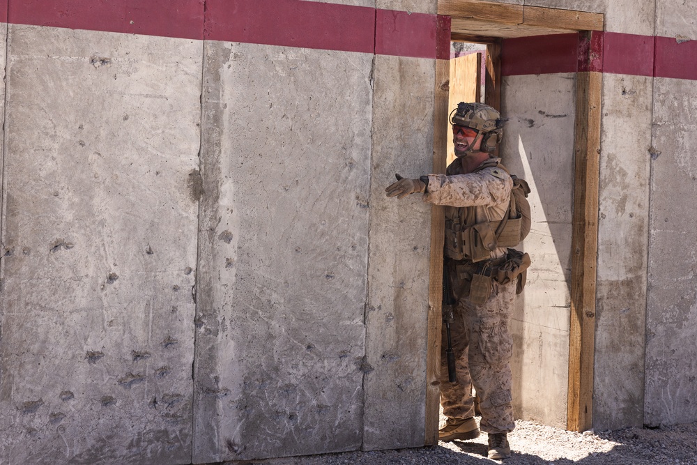 A soldier in desert camouflage uniform is positioned at a doorway in a concrete wall, appearing to be conducting a tactical entry or clearing exercise. The individual is wearing full combat gear including helmet, eye protection, body armor, and tactical vest. The setting appears to be a military training facility based on the constructed wooden door frame and painted concrete wall.