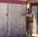 A soldier in desert camouflage uniform is positioned at a doorway in a concrete wall, appearing to be conducting a tactical entry or clearing exercise. The individual is wearing full combat gear including helmet, eye protection, body armor, and tactical vest. The setting appears to be a military training facility based on the constructed wooden door frame and painted concrete wall.