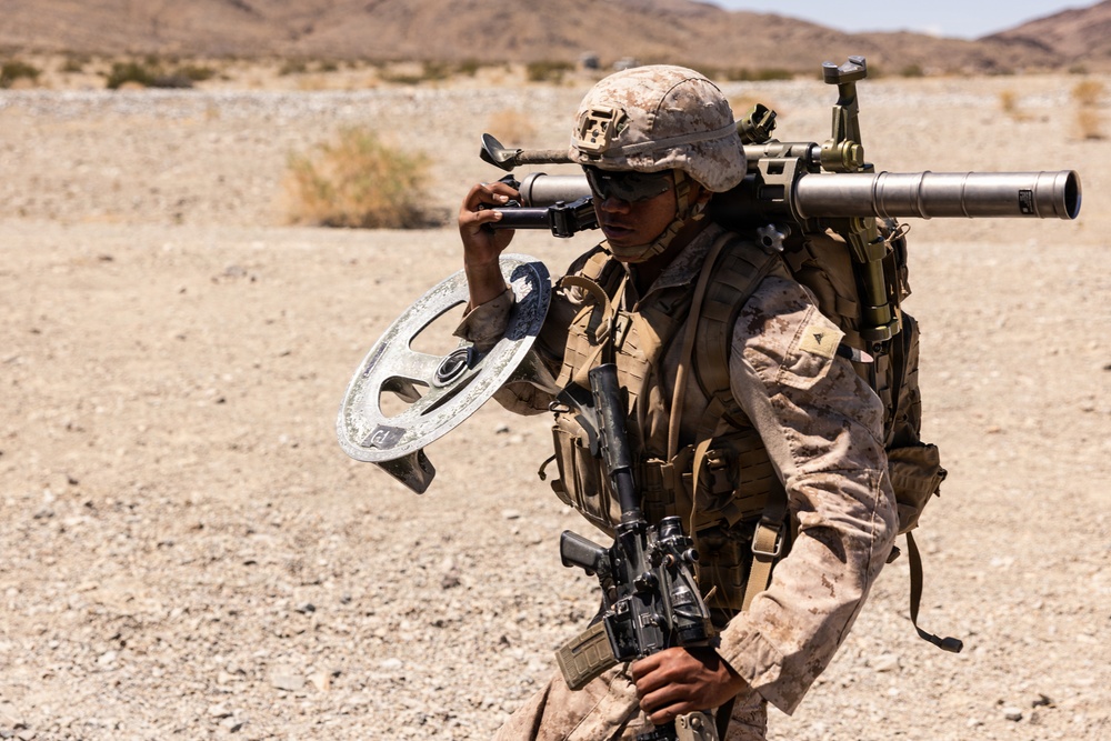 A U.S. Marine in desert MARPAT camouflage is carrying multiple pieces of equipment across an arid, desert terrain. The soldier is equipped with a metal detector, a shoulder-mounted weapon system (likely an AT4 or similar rocket launcher), and a slung assault rifle. The setting appears to be a training environment based on the dry, rocky landscape consistent with locations such as Twentynine Palms, California.
