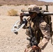 A U.S. Marine in desert MARPAT camouflage is carrying multiple pieces of equipment across an arid, desert terrain. The soldier is equipped with a metal detector, a shoulder-mounted weapon system (likely an AT4 or similar rocket launcher), and a slung assault rifle. The setting appears to be a training environment based on the dry, rocky landscape consistent with locations such as Twentynine Palms, California.