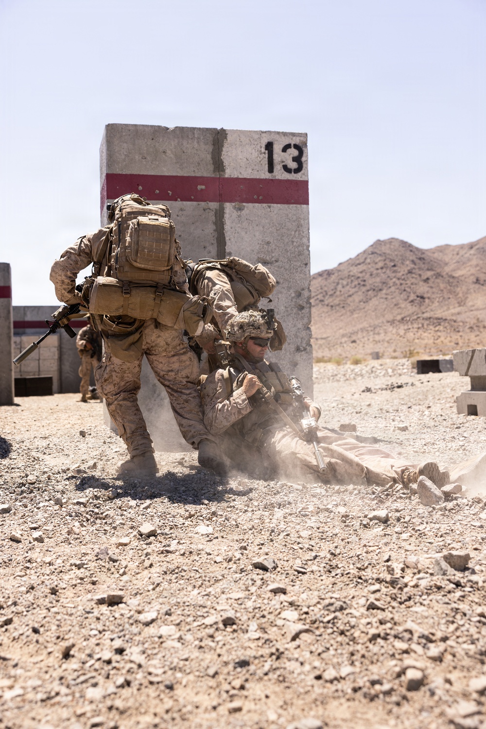 Two military personnel in desert camouflage uniforms are engaged in a casualty evacuation drill near a concrete barrier marked with the number '13' and a red stripe. One soldier is dragging another across rocky desert terrain, with dust being kicked up by the movement. A third service member is visible in the background, and the setting appears to be a military training range in an arid desert environment.