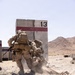 Two military personnel in desert camouflage uniforms are engaged in a casualty evacuation drill near a concrete barrier marked with the number '13' and a red stripe. One soldier is dragging another across rocky desert terrain, with dust being kicked up by the movement. A third service member is visible in the background, and the setting appears to be a military training range in an arid desert environment.