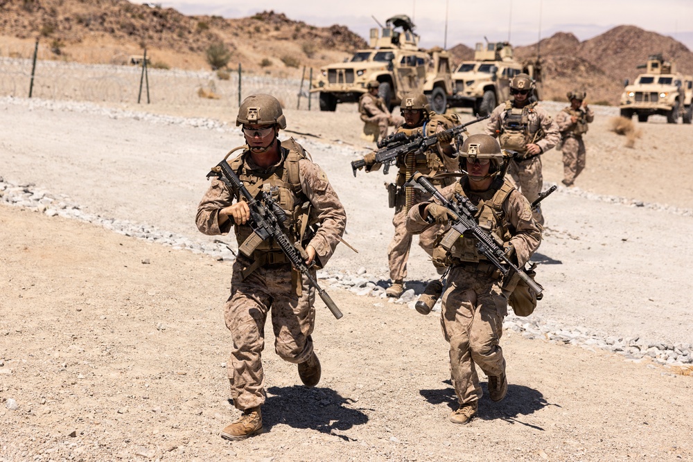 A group of military personnel in desert camouflage uniforms are moving on foot along a gravel road in an arid, desert environment. They are carrying assault rifles and wearing full combat gear including helmets, body armor, and tactical vests. Several tan military vehicles, including MRAPs and Humvees, are visible in the background.