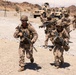 A group of military personnel in desert camouflage uniforms are moving on foot along a gravel road in an arid, desert environment. They are carrying assault rifles and wearing full combat gear including helmets, body armor, and tactical vests. Several tan military vehicles, including MRAPs and Humvees, are visible in the background.
