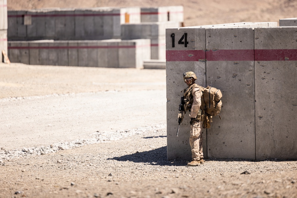 A fully equipped soldier in desert camouflage uniform stands against a large numbered concrete barrier (marked '14') with a red stripe. The setting appears to be a military training range or forward operating base in an arid, desert environment. The soldier is in a tactical posture, holding a rifle and wearing full combat gear including a helmet, body armor, and a large rucksack.