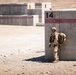 A fully equipped soldier in desert camouflage uniform stands against a large numbered concrete barrier (marked '14') with a red stripe. The setting appears to be a military training range or forward operating base in an arid, desert environment. The soldier is in a tactical posture, holding a rifle and wearing full combat gear including a helmet, body armor, and a large rucksack.