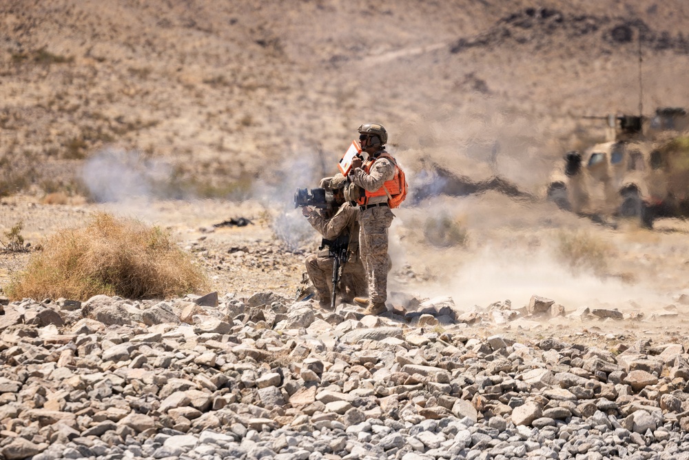Two military personnel in desert camouflage uniforms are operating in a rocky, arid terrain with smoke visible in the background. One soldier is wearing a bright orange safety vest and appears to be communicating via radio while the other operates what appears to be a crew-served weapon system. A military tactical vehicle (likely an MRAP or JLTV) is visible in the background to the right.
