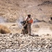 Two military personnel in desert camouflage uniforms are operating in a rocky, arid terrain with smoke visible in the background. One soldier is wearing a bright orange safety vest and appears to be communicating via radio while the other operates what appears to be a crew-served weapon system. A military tactical vehicle (likely an MRAP or JLTV) is visible in the background to the right.
