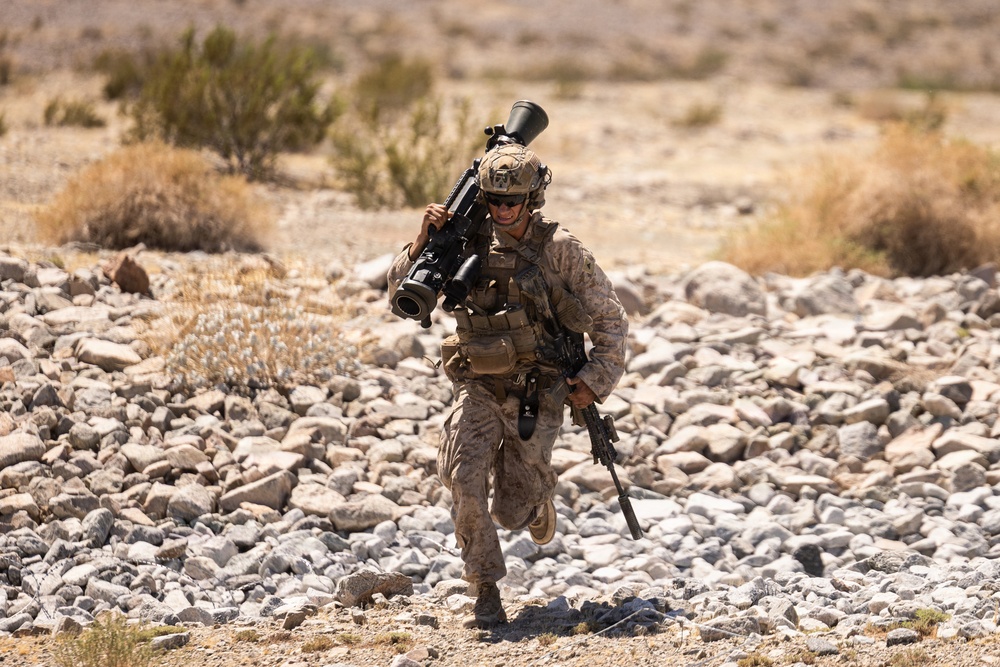 A U.S. Marine in desert MARPAT camouflage is running across rocky terrain in an arid environment. The soldier is carrying a shoulder-mounted weapon system (appears to be an M3E1 MAAWS/Carl Gustaf recoilless rifle) on his left shoulder and an M4/M16-pattern rifle in his right hand. The setting appears to be a desert training environment, likely in the American Southwest.