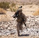 A U.S. Marine in desert MARPAT camouflage is running across rocky terrain in an arid environment. The soldier is carrying a shoulder-mounted weapon system (appears to be an M3E1 MAAWS/Carl Gustaf recoilless rifle) on his left shoulder and an M4/M16-pattern rifle in his right hand. The setting appears to be a desert training environment, likely in the American Southwest.