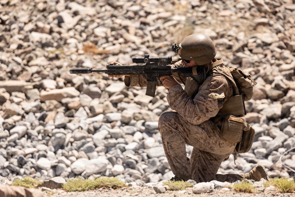 A soldier in full combat gear is kneeling on rocky terrain and aiming a rifle with an optical scope. The individual is wearing desert camouflage uniform, a tactical helmet, body armor, and a backpack. The setting appears to be an arid, rocky environment consistent with a training exercise or operational deployment.