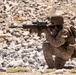 A soldier in full combat gear is kneeling on rocky terrain and aiming a rifle with an optical scope. The individual is wearing desert camouflage uniform, a tactical helmet, body armor, and a backpack. The setting appears to be an arid, rocky environment consistent with a training exercise or operational deployment.
