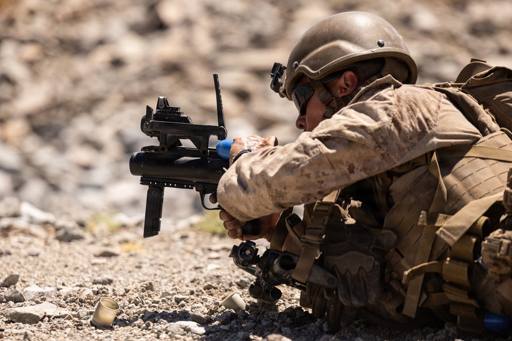 A soldier in desert camouflage uniform is in a prone/low firing position aiming a standalone grenade launcher, likely an M320 or similar weapon system. The soldier is equipped with full combat gear including a ballistic helmet with NVG mount, tactical vest, and backpack. Spent casings are visible on the rocky ground, suggesting active or recent firing.