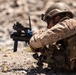 A soldier in desert camouflage uniform is in a prone/low firing position aiming a standalone grenade launcher, likely an M320 or similar weapon system. The soldier is equipped with full combat gear including a ballistic helmet with NVG mount, tactical vest, and backpack. Spent casings are visible on the rocky ground, suggesting active or recent firing.