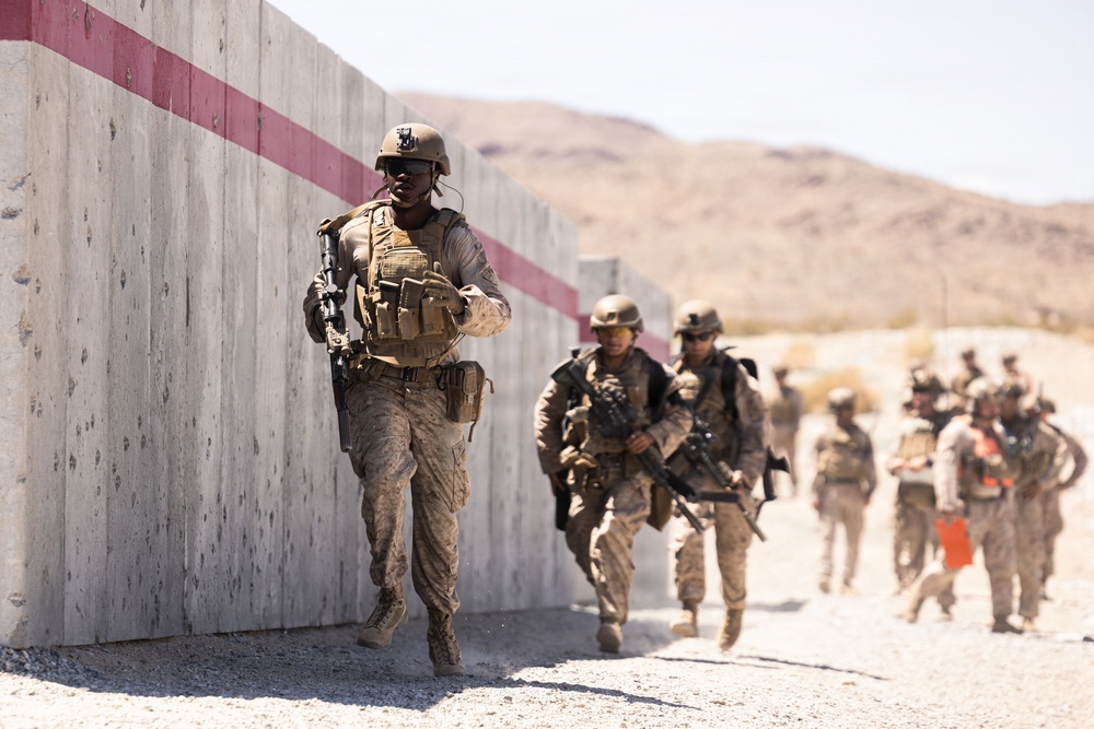 A group of military personnel in desert camouflage uniforms are conducting what appears to be a tactical training exercise in an arid environment. They are moving along a concrete wall with a red stripe, carrying rifles and wearing full combat gear including helmets, body armor, and tactical vests. The setting appears to be a military training facility based on the constructed concrete barriers and desert terrain.
