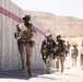 A group of military personnel in desert camouflage uniforms are conducting what appears to be a tactical training exercise in an arid environment. They are moving along a concrete wall with a red stripe, carrying rifles and wearing full combat gear including helmets, body armor, and tactical vests. The setting appears to be a military training facility based on the constructed concrete barriers and desert terrain.