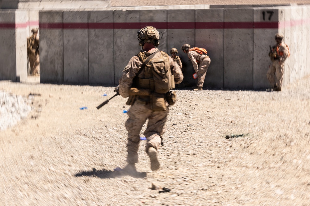 Military personnel in desert camouflage uniforms are conducting what appears to be a training exercise at a range or tactical training facility. A central figure is running toward a concrete wall structure marked with the number '17' and a red stripe. Multiple personnel are visible at various positions around the structure, some wearing orange safety vests indicating a supervised training environment.