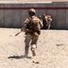 Military personnel in desert camouflage uniforms are conducting what appears to be a training exercise at a range or tactical training facility. A central figure is running toward a concrete wall structure marked with the number '17' and a red stripe. Multiple personnel are visible at various positions around the structure, some wearing orange safety vests indicating a supervised training environment.