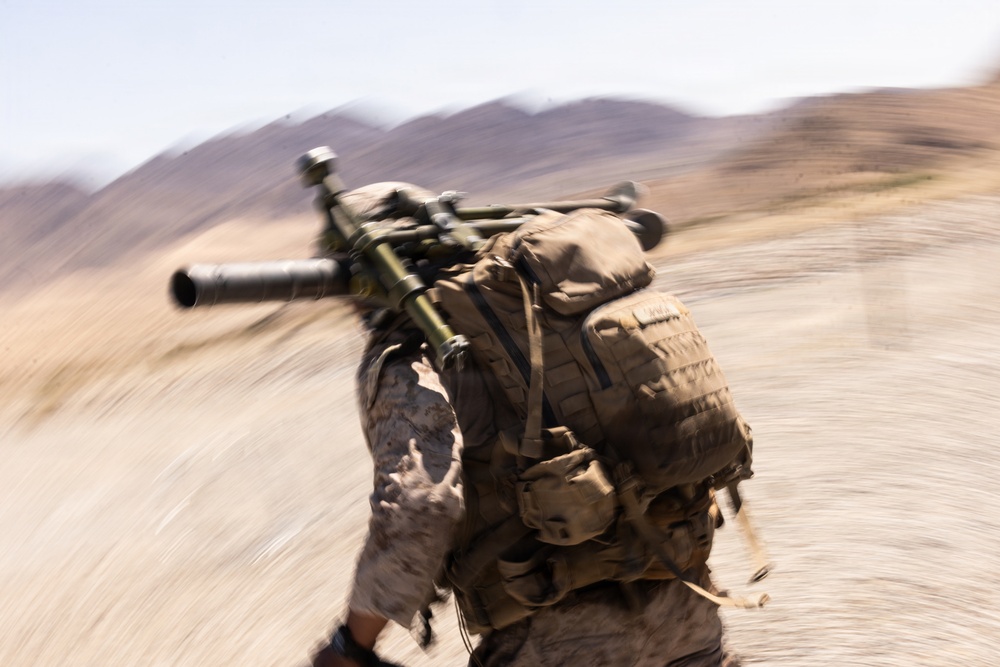 A military service member in desert camouflage uniform is captured in motion, running or moving rapidly through an arid, desert terrain. The soldier is carrying a large crew-served weapon system, likely a mortar or recoilless rifle, across their back along with a tactical backpack. The image is intentionally or incidentally motion-blurred, conveying a sense of urgency and rapid movement.
