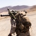 A military service member in desert camouflage uniform is captured in motion, running or moving rapidly through an arid, desert terrain. The soldier is carrying a large crew-served weapon system, likely a mortar or recoilless rifle, across their back along with a tactical backpack. The image is intentionally or incidentally motion-blurred, conveying a sense of urgency and rapid movement.