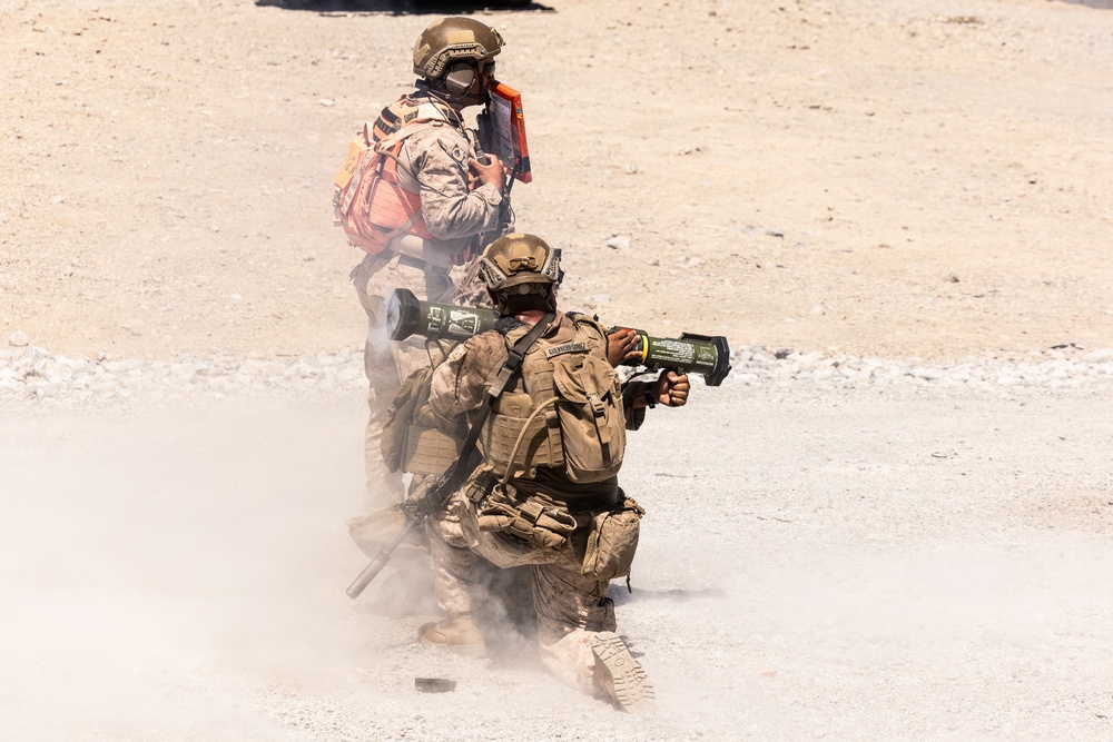 Two military personnel in desert camouflage uniforms are conducting a weapons firing exercise in an arid, open terrain environment. The foreground soldier is kneeling and firing what appears to be a shoulder-launched rocket or missile system, with visible backblast smoke and dust. A second soldier stands behind, wearing a red medical/safety pack and appearing to observe or coordinate the exercise.