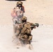 Two military personnel in desert camouflage uniforms are conducting a weapons firing exercise in an arid, open terrain environment. The foreground soldier is kneeling and firing what appears to be a shoulder-launched rocket or missile system, with visible backblast smoke and dust. A second soldier stands behind, wearing a red medical/safety pack and appearing to observe or coordinate the exercise.