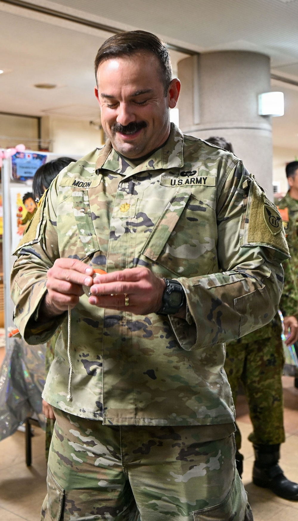 A U.S. Army Major (identified by gold oak leaf insignia) named 'Moon' is smiling and examining a small object in his hands while in what appears to be an indoor facility. He is wearing Operational Camouflage Pattern (OCP) uniform with U.S. Army tape, a name tape reading 'MOON', parachutist badge, and a unit patch on his left shoulder. Other personnel in what appear to be Japanese Self-Defense Force uniforms are visible in the background.