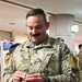 A U.S. Army Major (identified by gold oak leaf insignia) named 'Moon' is smiling and examining a small object in his hands while in what appears to be an indoor facility. He is wearing Operational Camouflage Pattern (OCP) uniform with U.S. Army tape, a name tape reading 'MOON', parachutist badge, and a unit patch on his left shoulder. Other personnel in what appear to be Japanese Self-Defense Force uniforms are visible in the background.